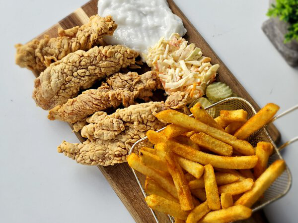 Crispy Chicken Strips mit Pommes, Slaw und Dip auf einem Holzbrett serviert.