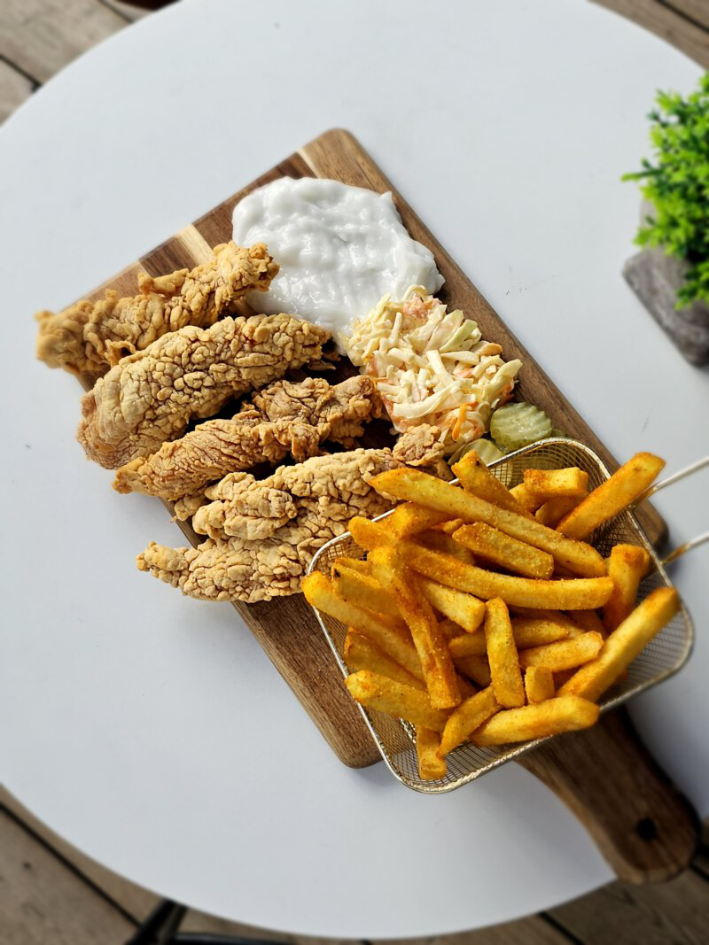 Crispy Chicken Strips mit Pommes, Slaw und Dip auf einem Holzbrett serviert.